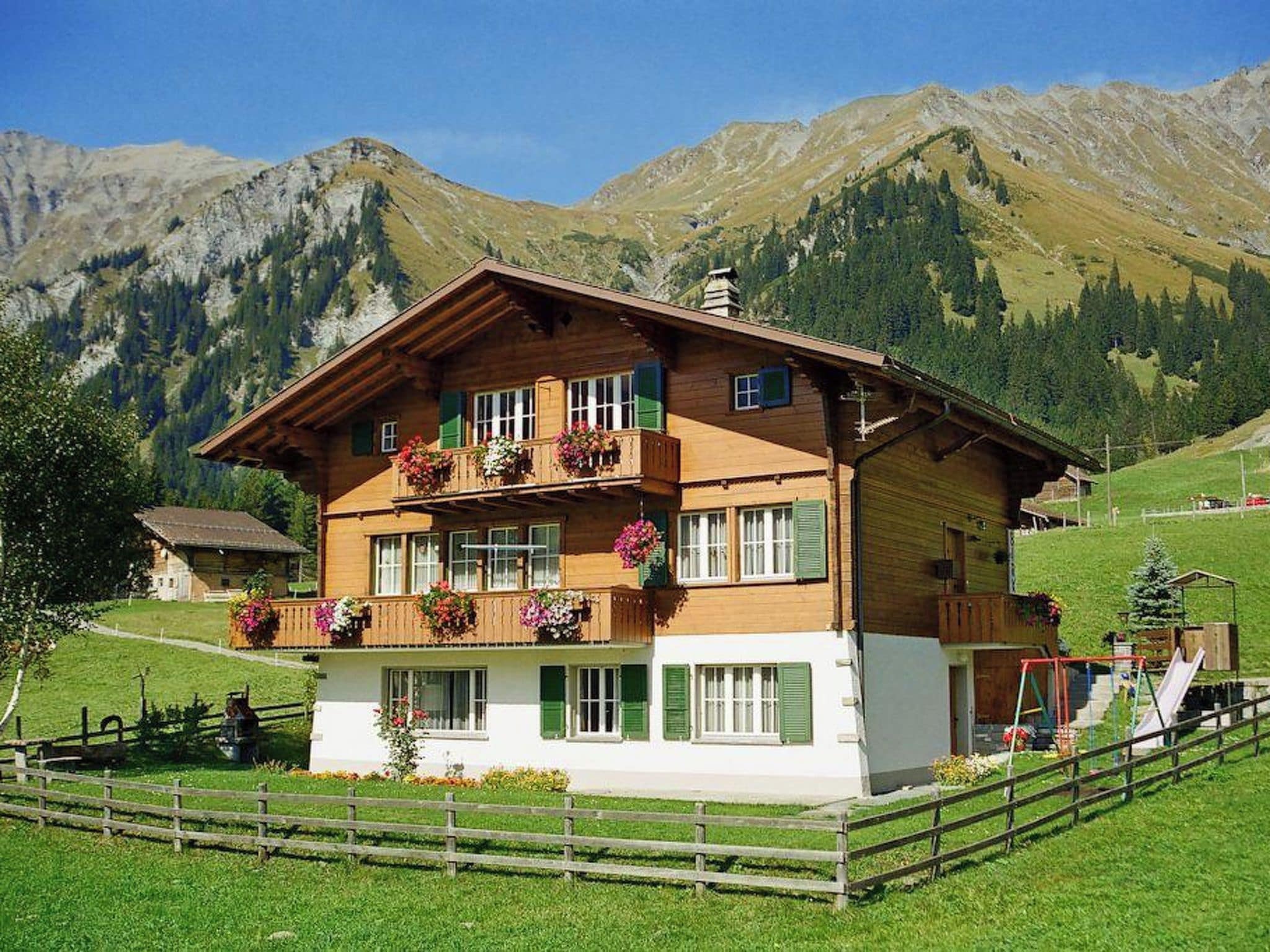 Un chalet à deux étages doté d’un balcon, entouré d’une clôture en bois et offrant une vue sur les montagnes.