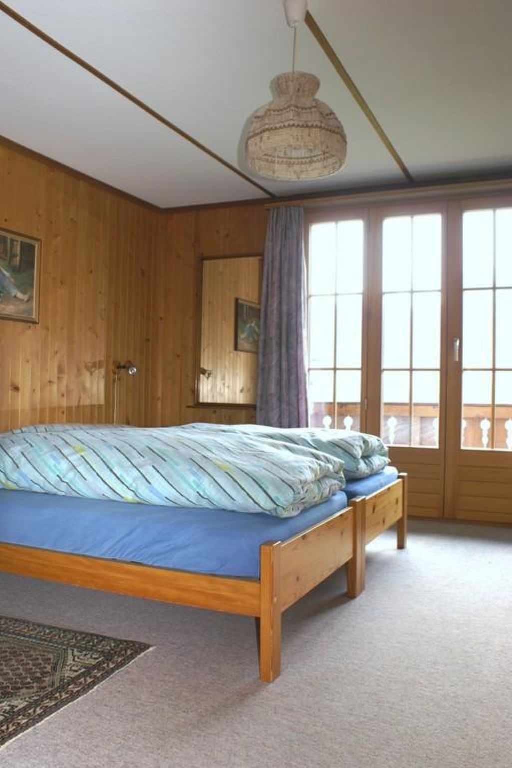Una habitación con una cama de madera, una lámpara colgante y un amplio ventanal con cortinas.