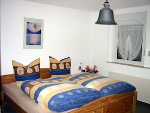 A bedroom with a wooden bed, a painting on the wall, a window with curtains, and a hanging lamp.