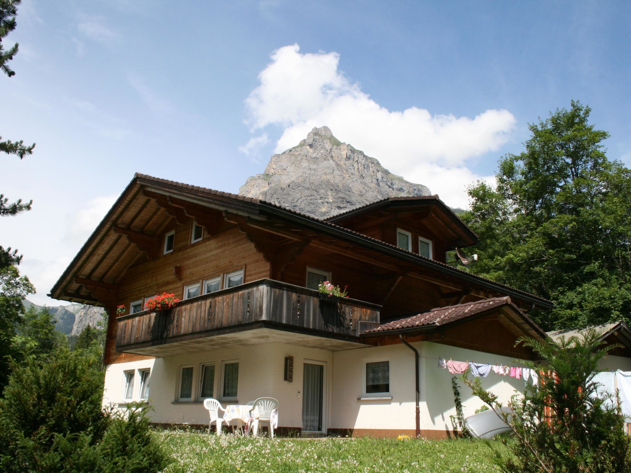 A two-story wooden chalet with a balcony, surrounded by greenery and a mountain backdrop.