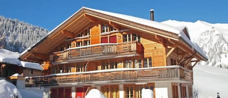 A multi-story wooden chalet with balconies, set in a snowy mountainous area.