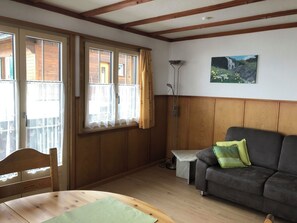 Ein Zimmer mit Holzboden, einem grauen Sofa mit einem grünen Kissen, einem Holzstuhl, einem kleinen Tisch und einem Bild an der Wand.