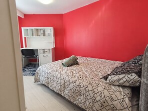 A bedroom with a red wall, a bed with patterned bedding, a desk with a chair, and a wardrobe. - The Family Home (Chartres)