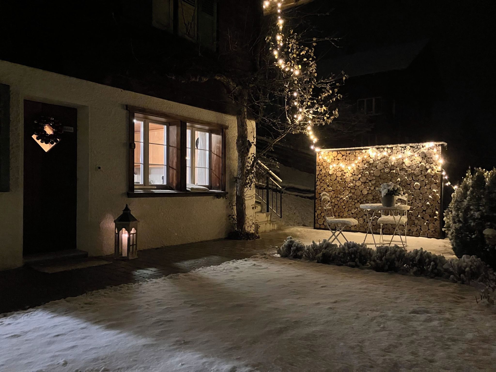 A snow-covered courtyard with a wooden bench and a table, a tree with lights, and a building with windows.