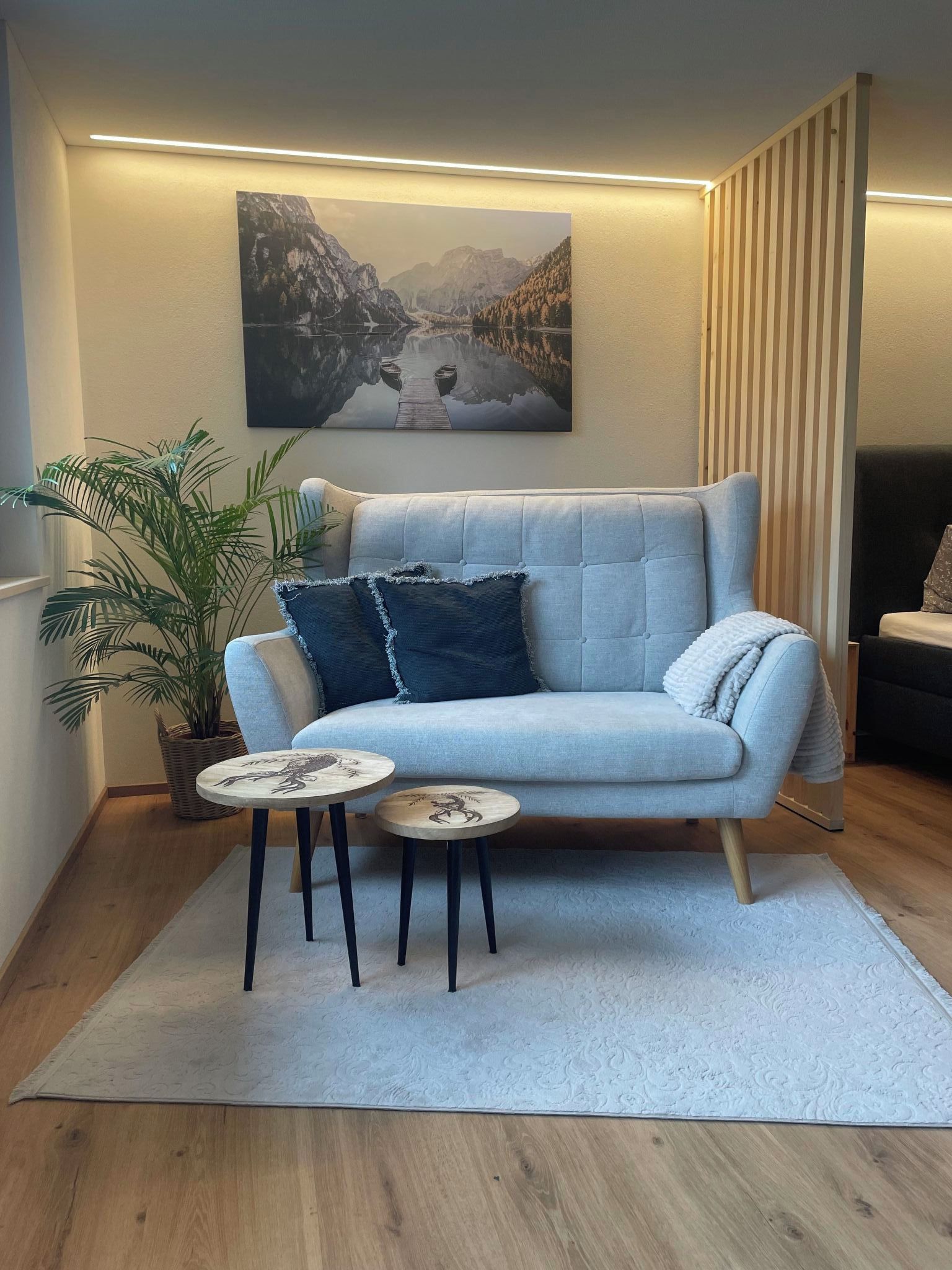 Un salon avec un canapé bleu clair, une petite table ronde surmontée d’un élément décoratif, une grande plante en pot et un tableau représentant un paysage montagneux.