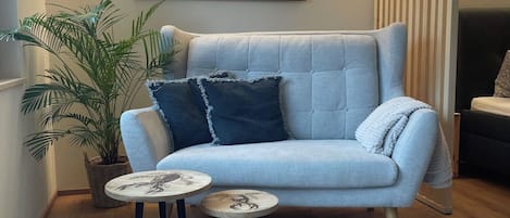Un salon avec un canapé bleu clair, une petite table ronde surmontée d’un élément décoratif, une grande plante en pot et un tableau représentant un paysage montagneux.