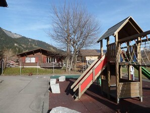 Ein Spielplatz mit Rutsche und Kletterbereich, umgeben von Gebäuden und einem Berg im Hintergrund.