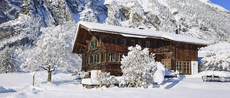 A wooden chalet with a snow-covered roof, surrounded by snow-laden trees and mountains.