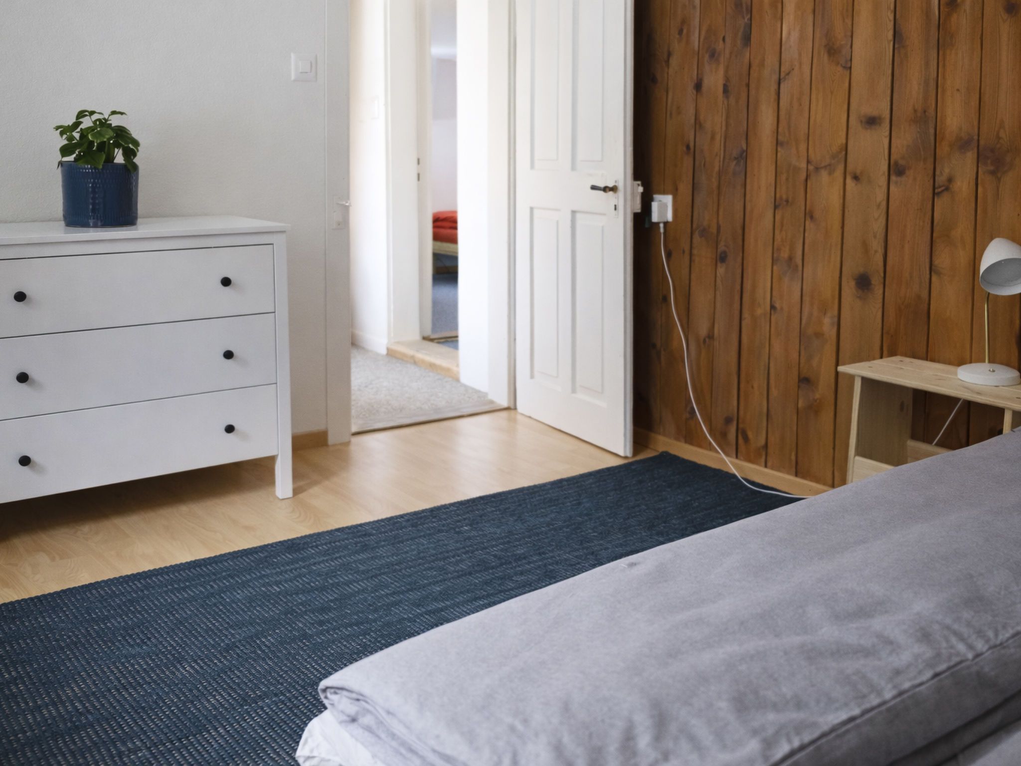 A bedroom with a bed, a dresser, a nightstand, a lamp, and a wooden wall.