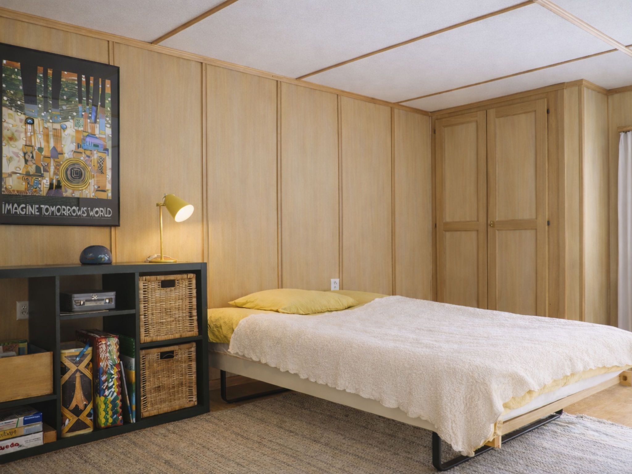 A bedroom with a single bed, a nightstand, a lamp, a bookshelf, and a framed picture on the wall.