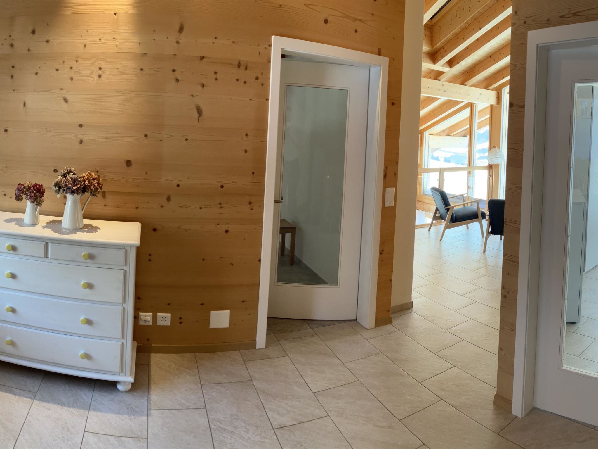 Ein Holzinterieur mit einer weißen Kommode, einer Tür, die auf einen Balkon führt, und Blick ins Freie.
