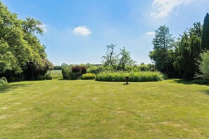 A well-maintained grassy area with a variety of trees and shrubs, a clear sky, and a distant view of a hill. - Village school from 1777 in West Lolland (Vesterborg)
