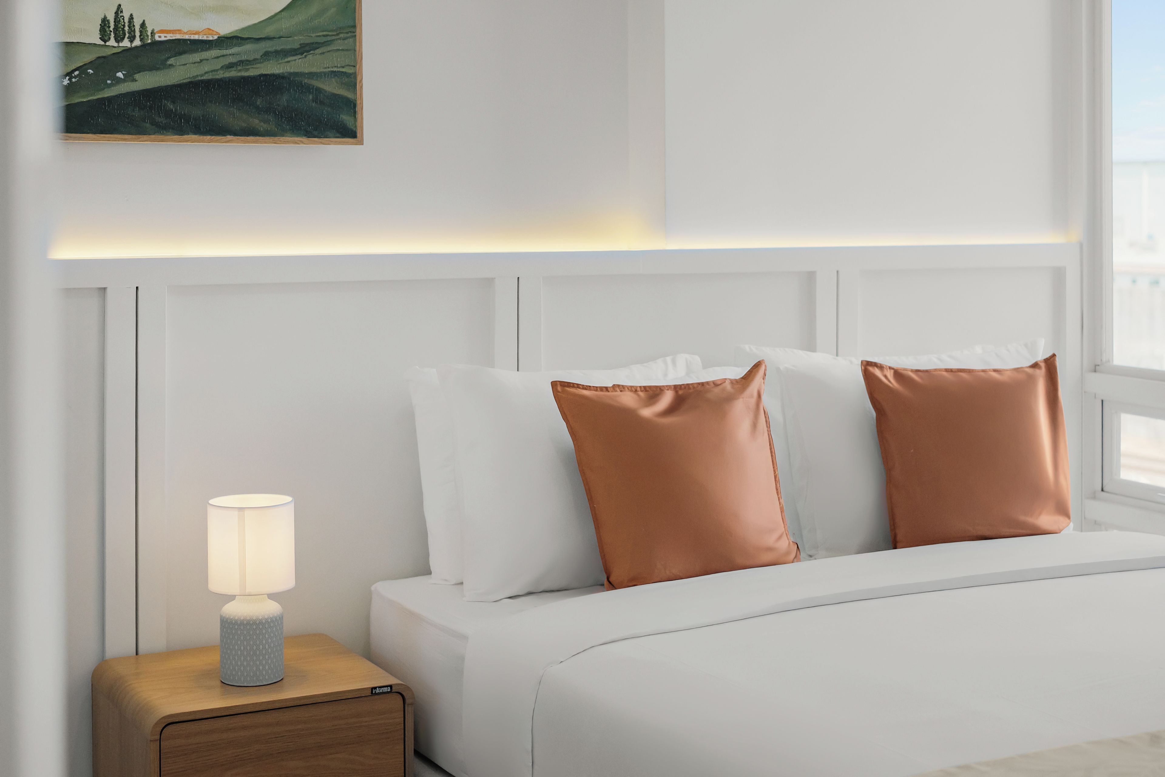 Una cama bien hecha con almohadas blancas y de cobre, una mesita de noche con una lámpara y un cuadro en la pared.