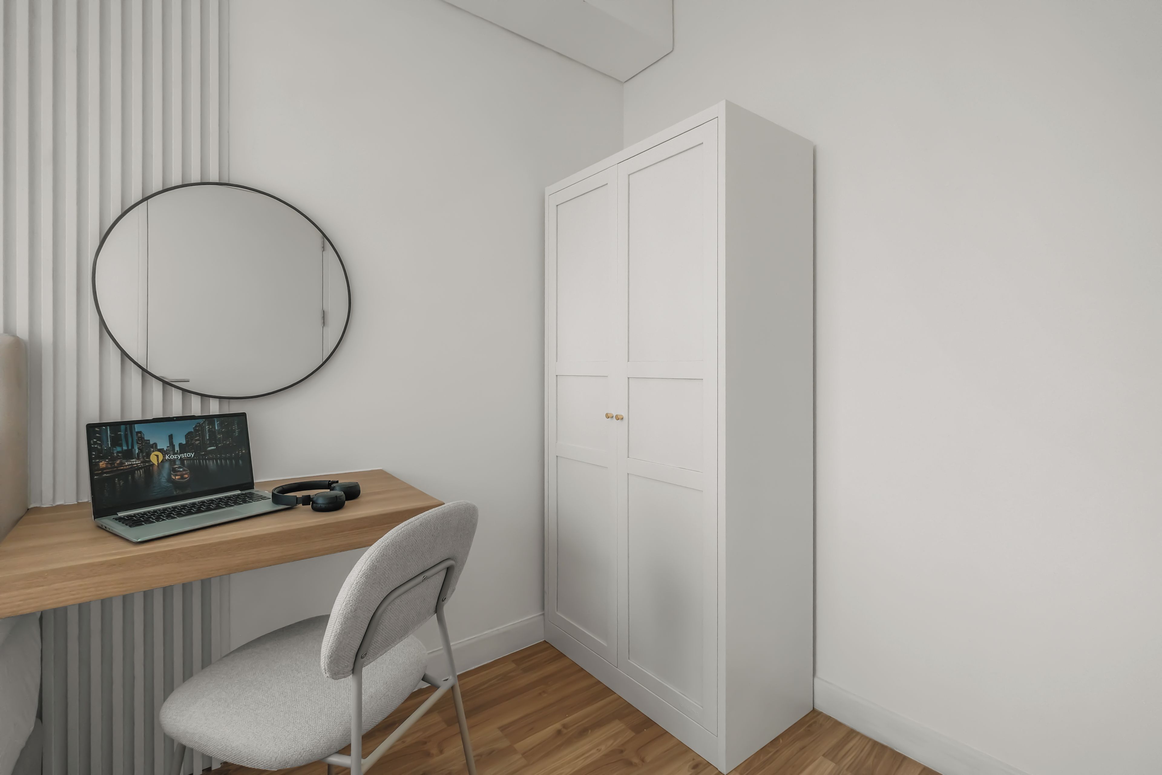 Une pièce comprenant un bureau en bois, un ordinateur portable, un casque audio, une chaise, un miroir rond et une armoire blanche.
