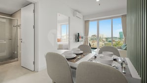 A modern dining area with a table set for four, a TV mounted on the wall, and a view of the cityscape through large windows. - Asriya by Kozystay - Paskal (Bandung)