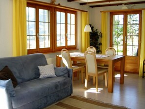 Ein Wohnzimmer mit einer Couch, einem Esstisch, Stühlen und großen Fenstern mit Vorhängen.