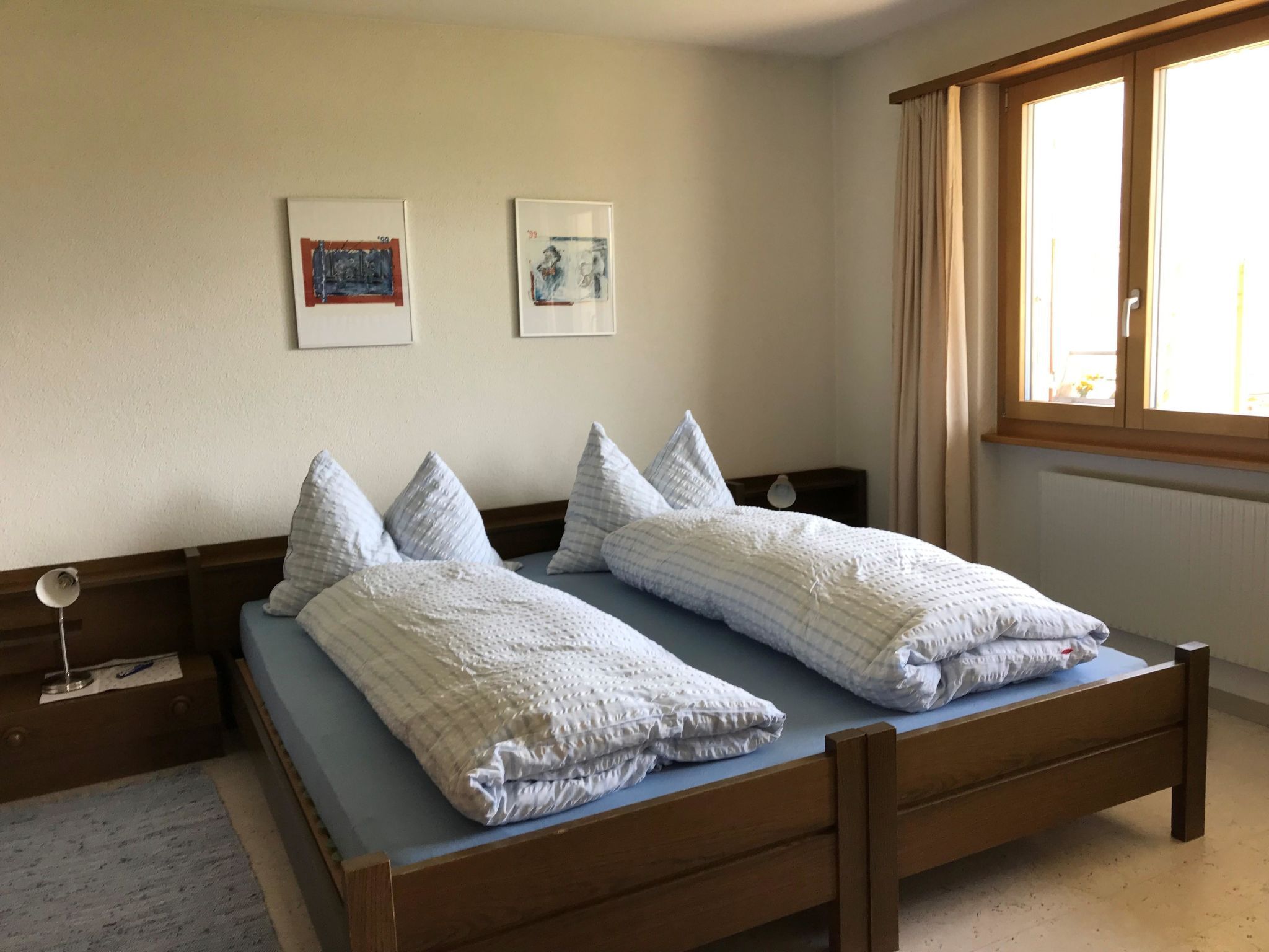 Uma cama de casal com estrutura de madeira e colchão azul, duas mesas de cabeceira com luminárias e duas obras de arte emolduradas na parede.