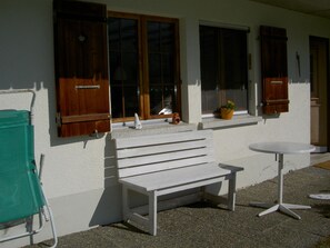 Eine weiße Bank und ein Tisch vor einem Gebäude mit hölzernen Fensterläden und einem Blumentopf am Fensterbrett.