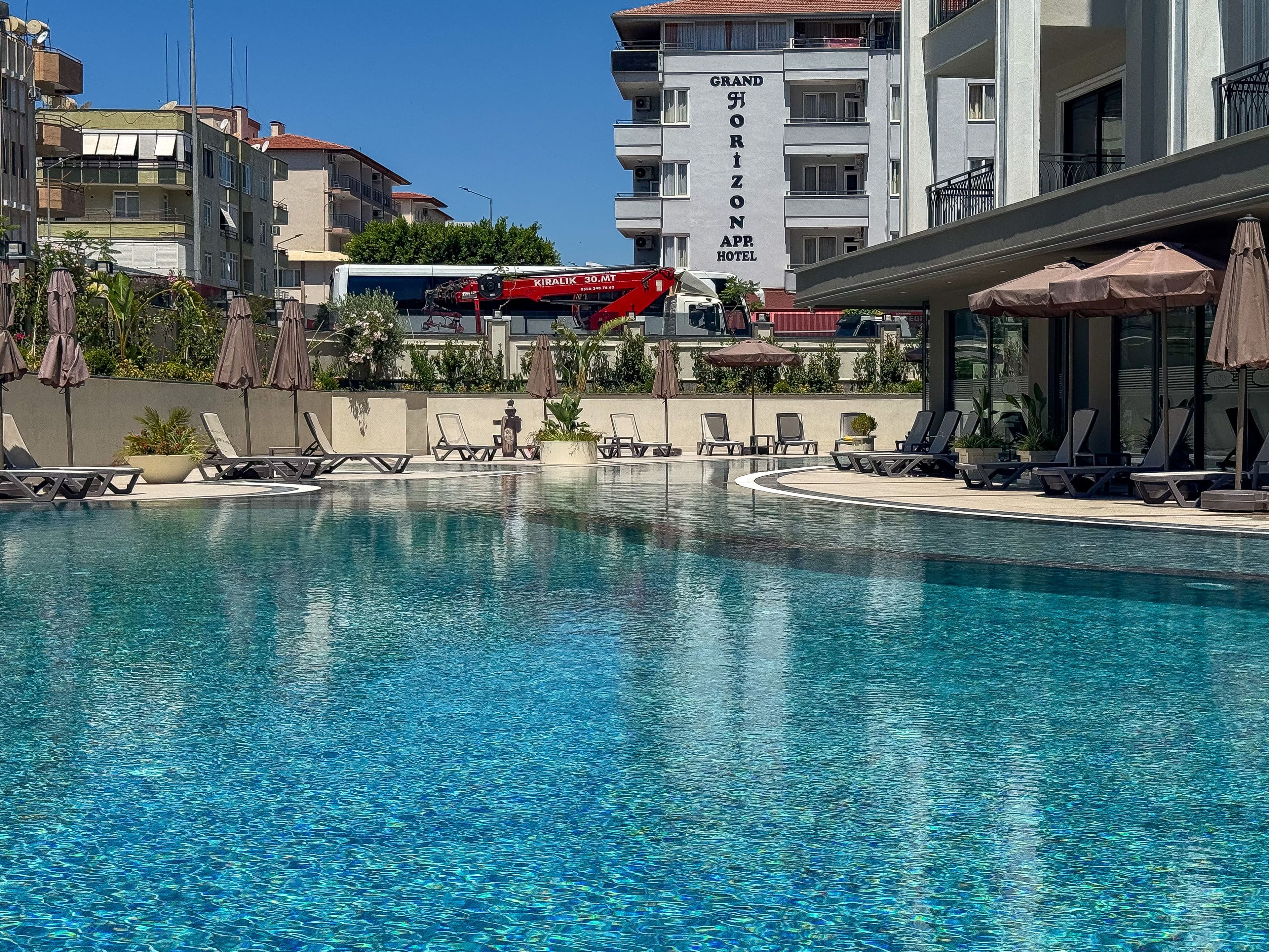 A swimming pool with lounge chairs and umbrellas, a building with a sign reading 'Grand Horizon App Hotel', and a red crane in the background.