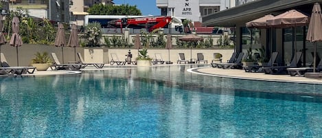 A swimming pool with lounge chairs and umbrellas, a building with a sign reading 'Grand Horizon App Hotel', and a red crane in the background.