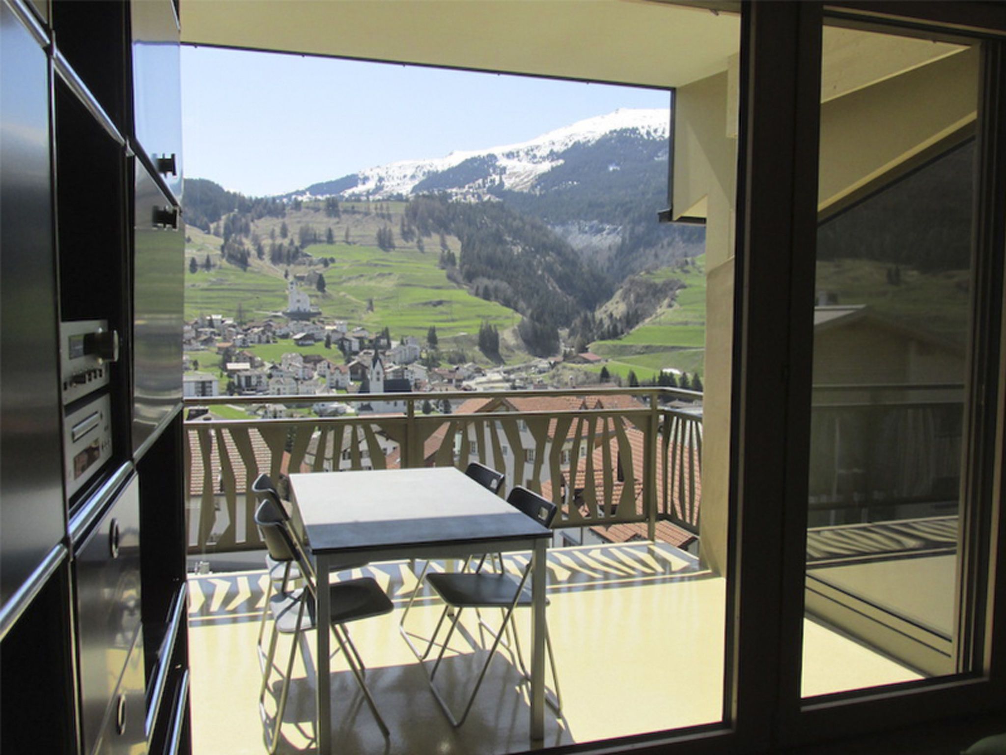 Un balcon avec une table et des chaises, d’où l’on a vue sur un village et des montagnes.