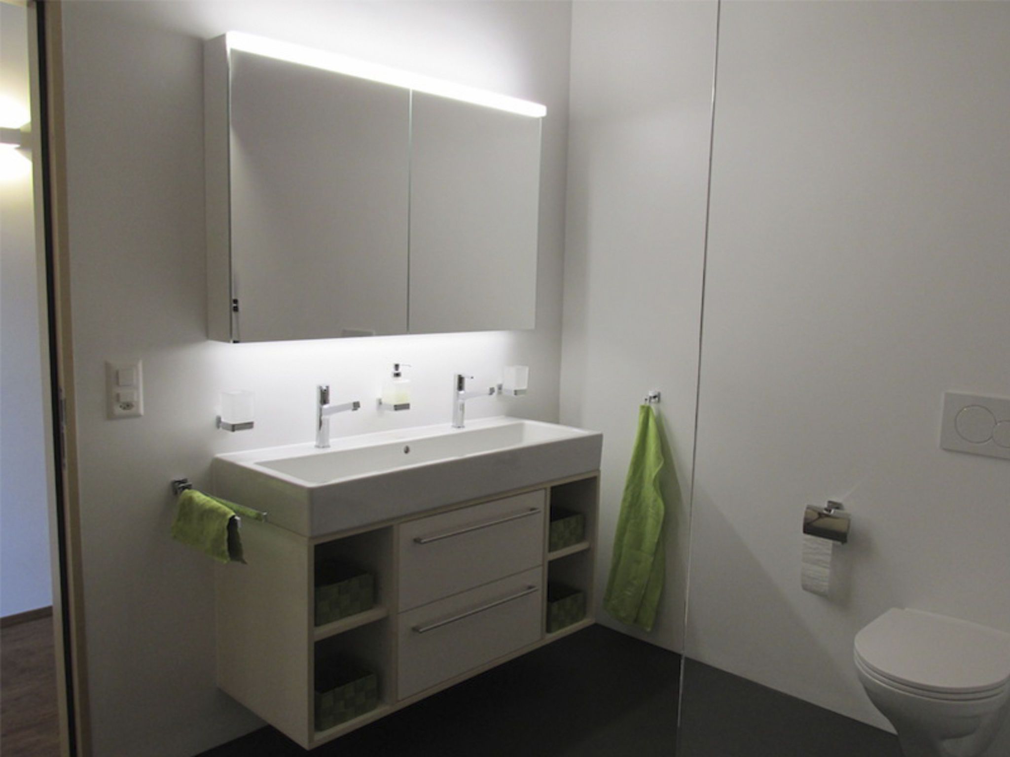 Ein modernes Badezimmer mit einem weißen Waschtisch, zwei Waschbecken, einem Spiegel und einer Toilette.