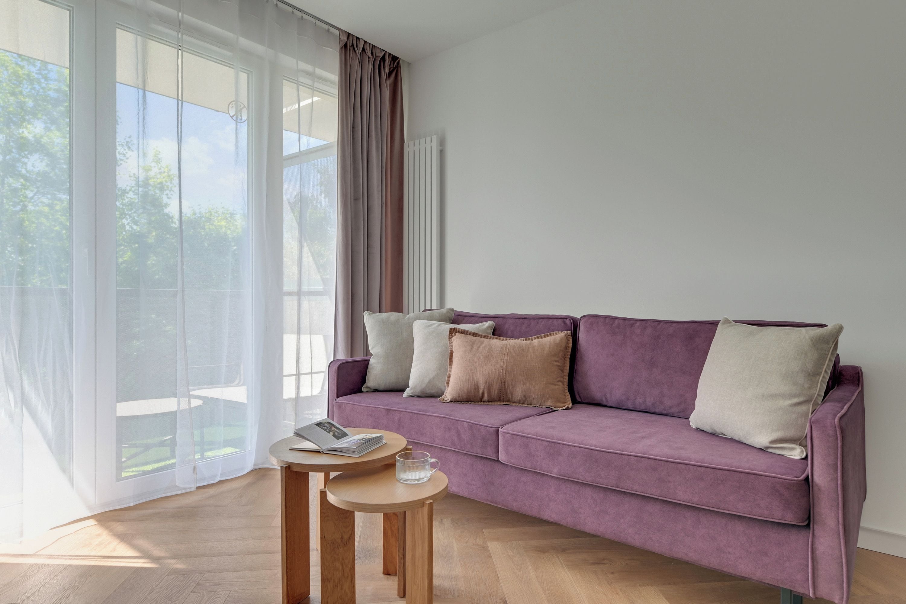 A modern living room with a purple sofa, a small wooden table, and a large window with curtains.