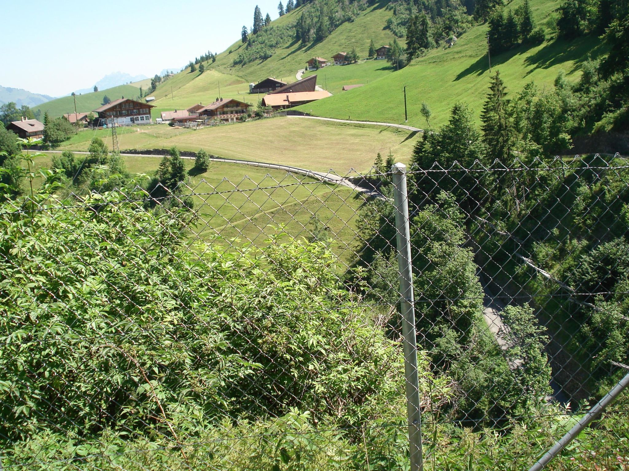 Un paysage rural avec des maisons, un espace délimité par une clôture et une chaîne de montagnes en arrière-plan.