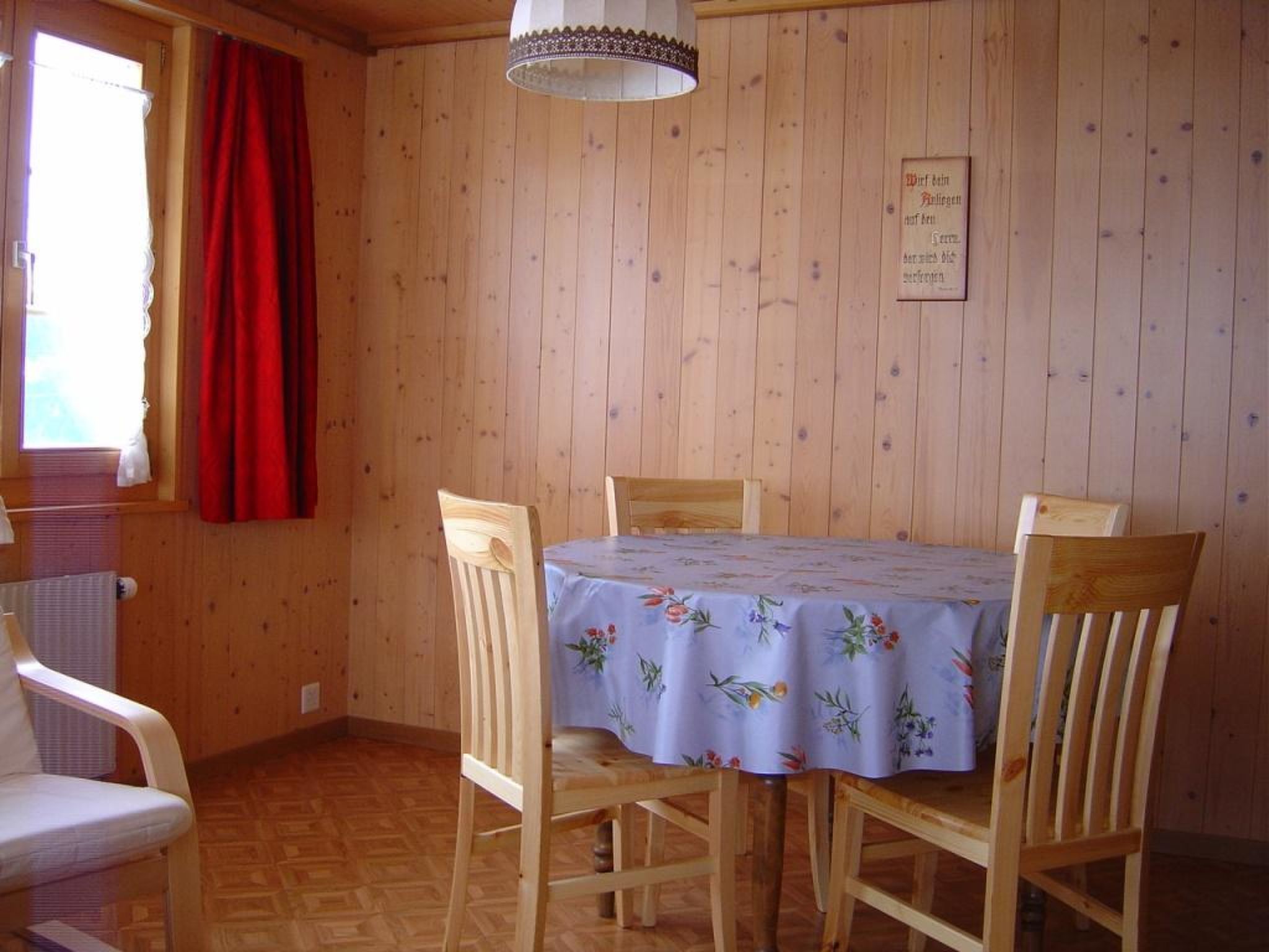 Une pièce aux murs lambrés, avec une table recouverte d’une nappe à motifs floraux, deux chaises en bois et un rideau rouge.