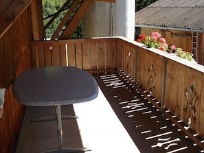 Un balcón de madera con una mesa y sillas de metal, una maceta con flores rosadas, y una vista de una casa con techo de zinc.