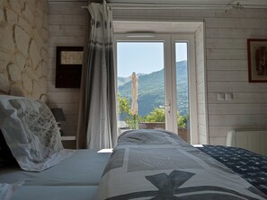 Een slaapkamer met een bed, een raam met gordijnen, uitzicht op bergen en een deur naar buiten.