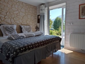 Een slaapkamer met een stenen muur, een bed met beddengoed met een patroon, een nachtkastje, een raam met gordijnen en uitzicht op groen en bergen.