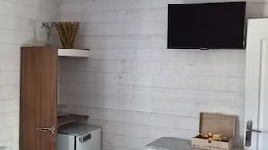 A small, compact room with a wooden wardrobe, a small refrigerator, a television mounted on the wall, and a box of items on the floor. - HARMONIE ROOM FOR 4 PEOPLE (Auris)
