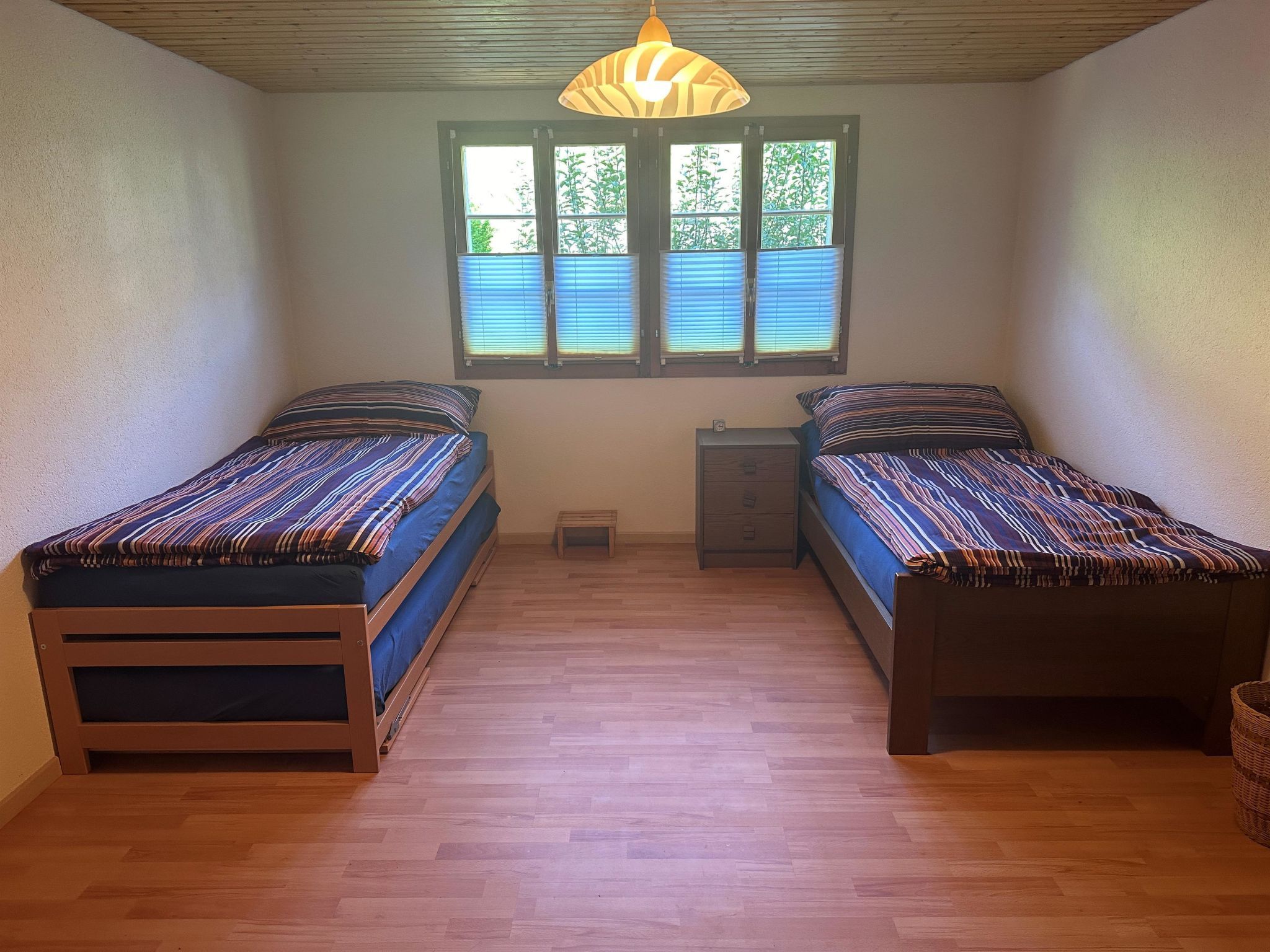 Une petite pièce bien éclairée, avec deux lits simples, un plancher en bois, une fenêtre avec des stores et un luminaire suspendu.