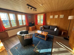 A cozy living room with a sofa, a coffee table, and a television. There are windows with curtains, a wooden ceiling, and a view of the outdoors. - Ferienwohnung Gehrihorn Appartement/Fewo, Dusche, WC, 2 Schlafräume by Interhome (Frutigen)