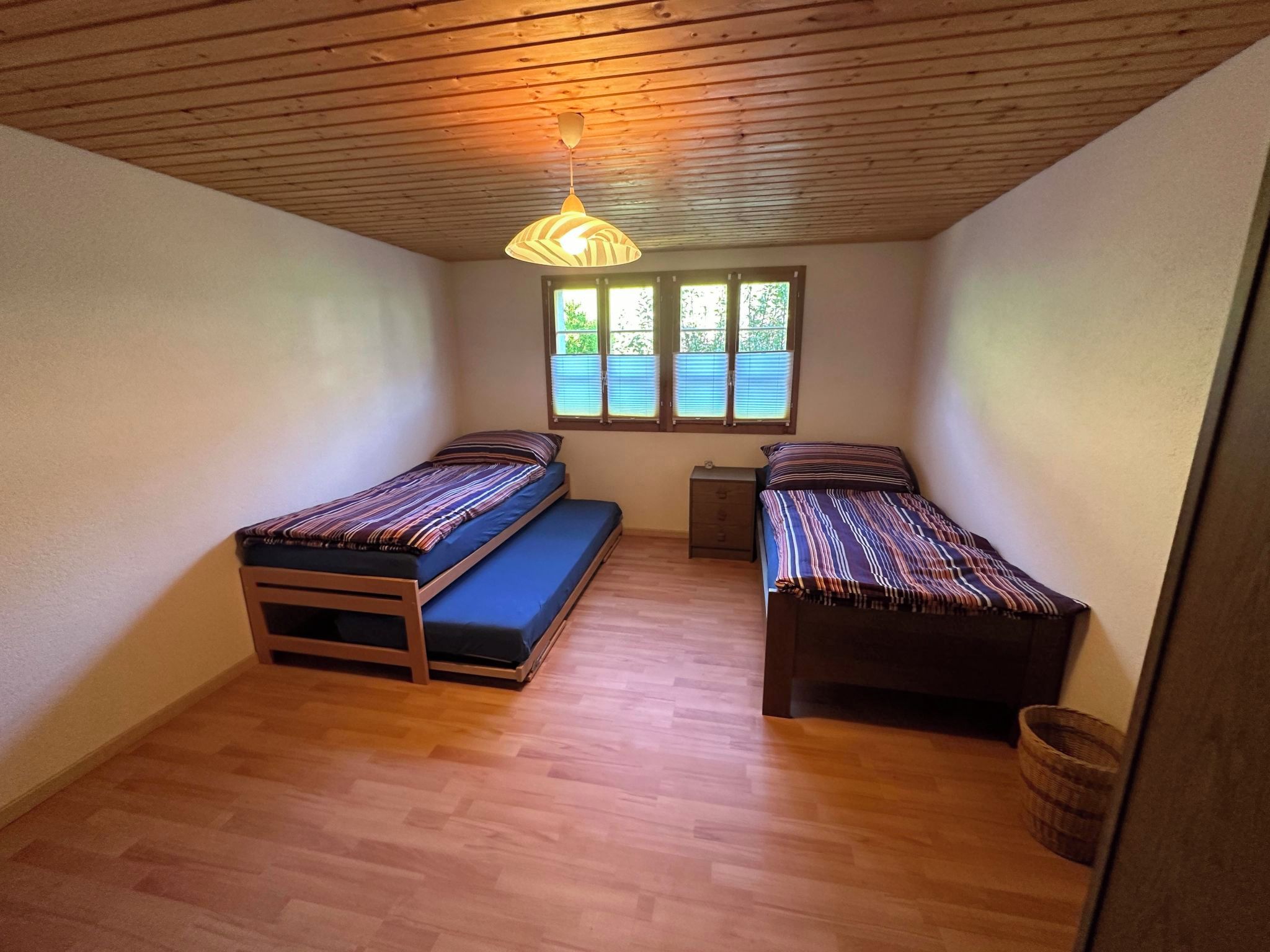 Quarto pequeno com duas camas de solteiro, teto de madeira, janela com persianas e luminária pendente.