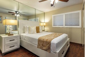 A bedroom with a bed, bedside tables, a ceiling fan, a mirror, and a window with blinds. - Maalaea Kai #220- Relaxing 2BR/2BA Oceanfront Maui Condo with Private Lanai, Stunning Ocean Views (Wailuku)
