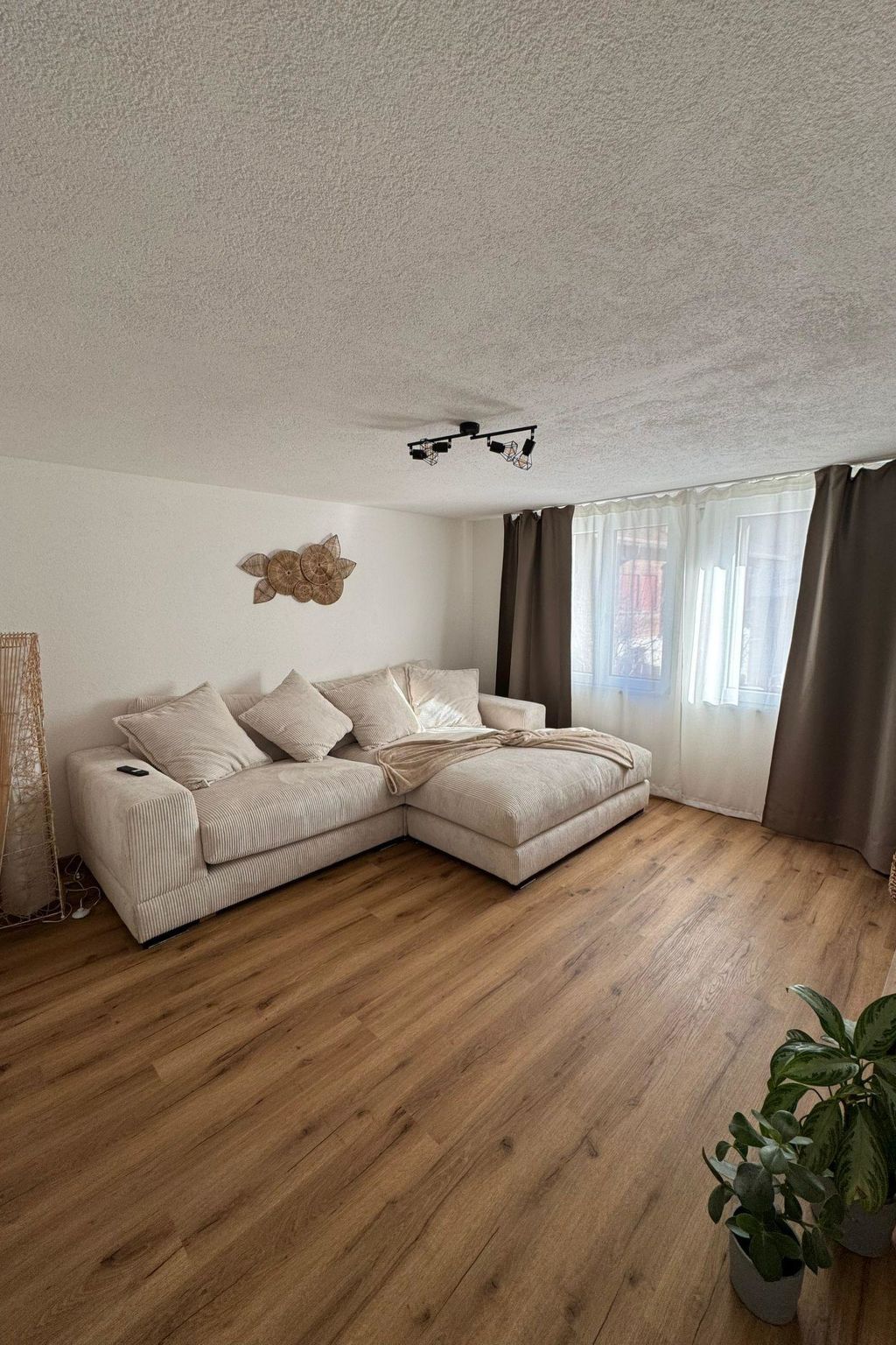 En stue med en beige hjørnesofa, trægulv, en loftlampe og en dekorativ genstand på væggen.