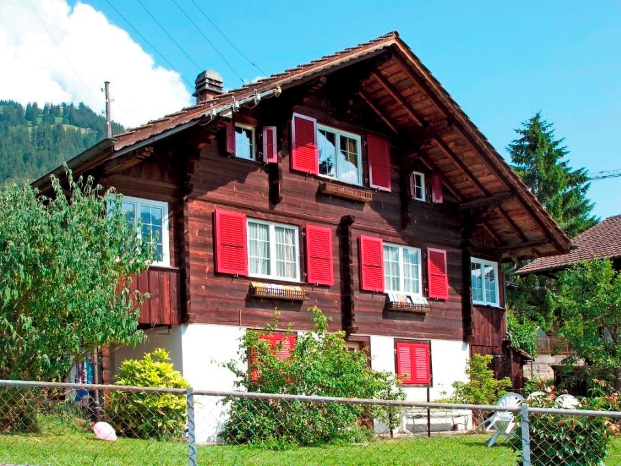 Una casa de madera de dos pisos con contraventanas rojas, rodeada de follaje y un cielo despejado.
