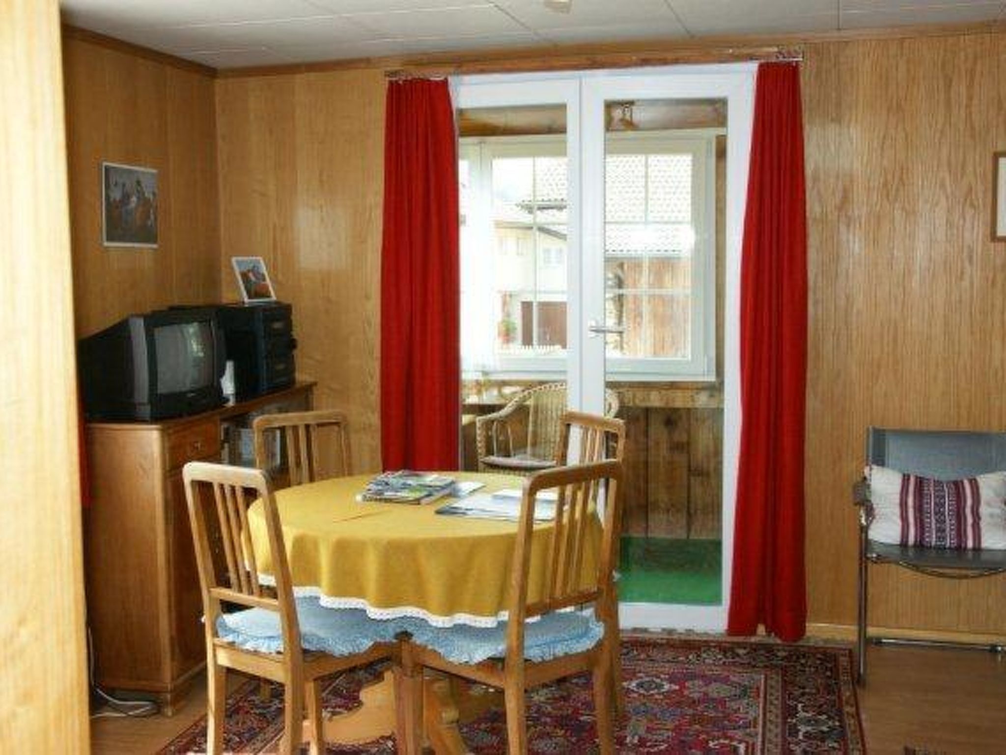 Un espace repas comprenant une table, des chaises, un téléviseur et une vue sur l’extérieur, visible à travers une porte vitrée.