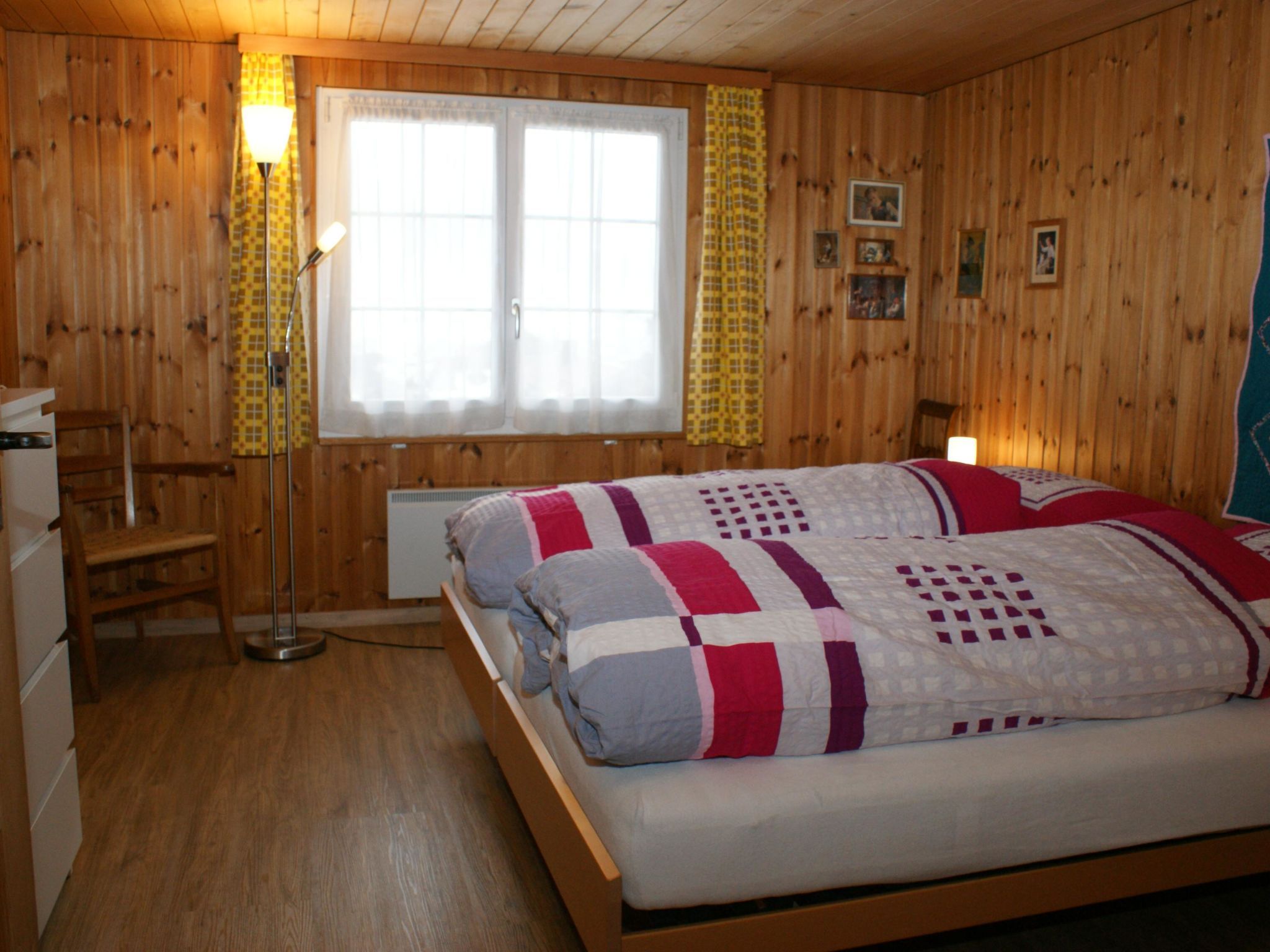 Ein Zimmer mit Holzwandverkleidung, einem Bett, einem Sessel, einer Stehlampe und einem Fenster mit Vorhängen.