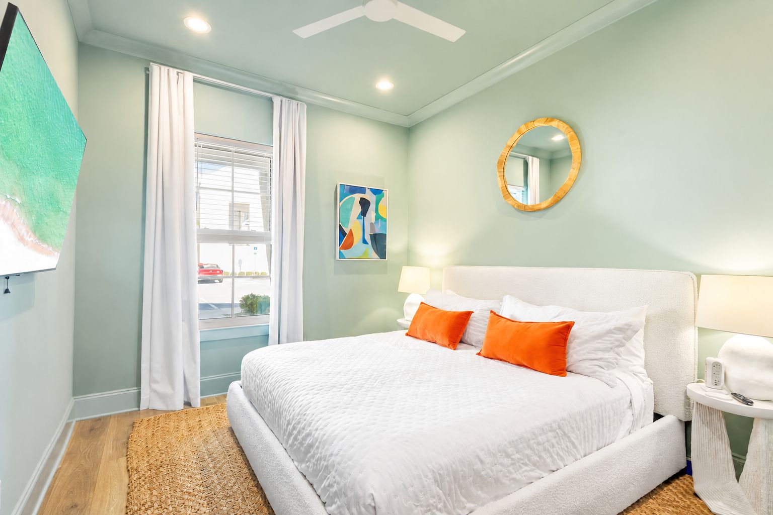 A hotel room with a bed, a bedside table, a mirror, a painting, and a window with curtains.