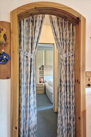 A doorway with patterned curtains leading to a bedroom with a bed, bedside table, lamp, and a wall-mounted shelf. - 4_3 Apartment WildentenNest Zellersee by Interhome (Zell am See)