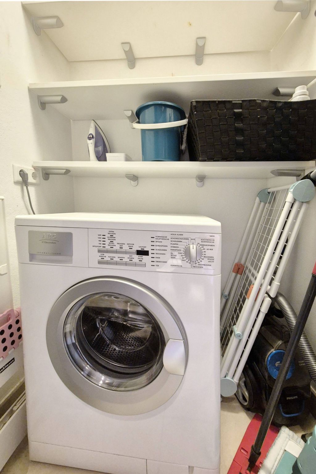 Een compacte wasmachine met een bedieningspaneel, een droogrek en een blauwe emmer op een plank erboven.