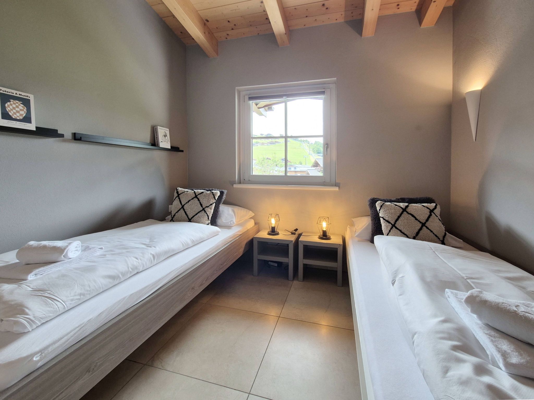 Een ruime, goed verlichte kamer met twee eenpersoonsbedden, een raam en een plank met boeken.