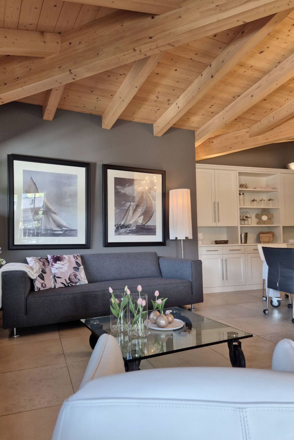 A modern living room with a grey sofa, a glass coffee table with decorative items, and a white armchair. There are two framed pictures on the wall, a lamp, and a kitchen area in the background.