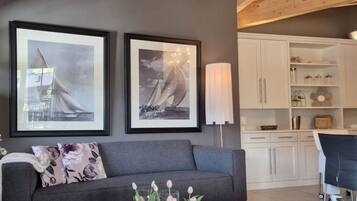 A modern living room with a grey sofa, a glass coffee table with decorative items, and a white armchair. There are two framed pictures on the wall, a lamp, and a kitchen area in the background.
