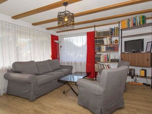 A living room with a grey sofa set, a glass coffee table, a bookshelf with books, a television, and a red curtain. - Kurhaus Appartement/Fewo, Dusche und Badewanne, 2 Schlafräume 3-Zimmerwohnung by Interhome (Adelboden)