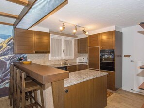 A modern kitchen with wooden cabinets, a granite countertop, and a built-in island with stools. - Kurhaus Appartement/Fewo, Dusche und Badewanne, 2 Schlafräume 3-Zimmerwohnung by Interhome (Adelboden)