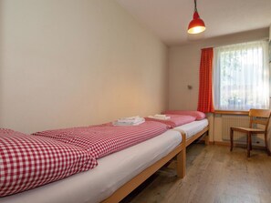 A room with two single beds, a wooden chair, a radiator, and a window with curtains. - Kurhaus Appartement/Fewo, Dusche und Badewanne, 2 Schlafräume 3-Zimmerwohnung by Interhome (Adelboden)